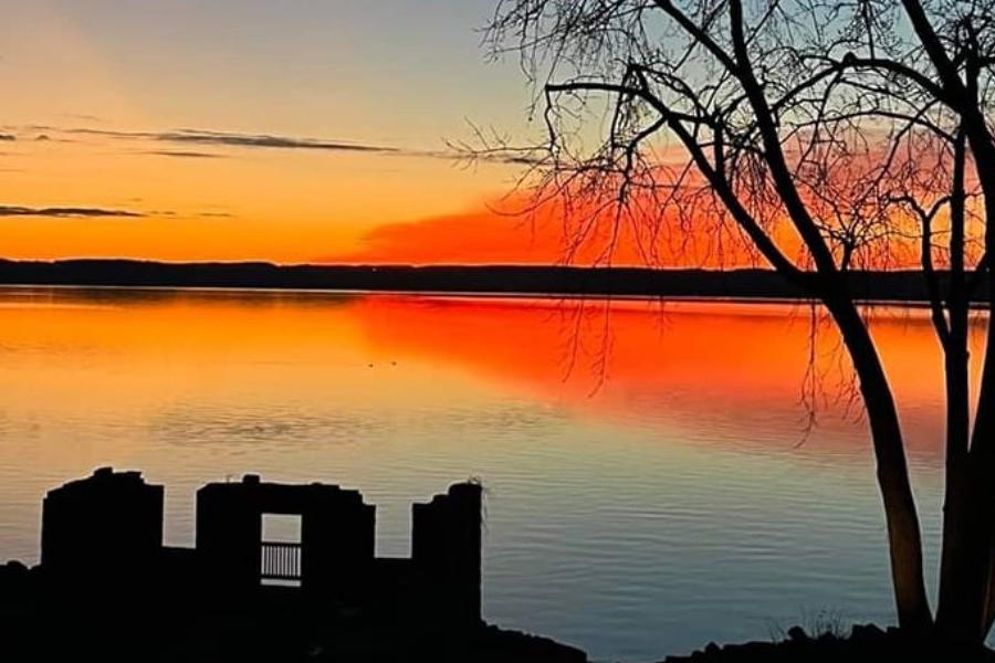 An orange and yellow sunset casts over a bed of water as a buildings remains and trees are seen in the foreground.